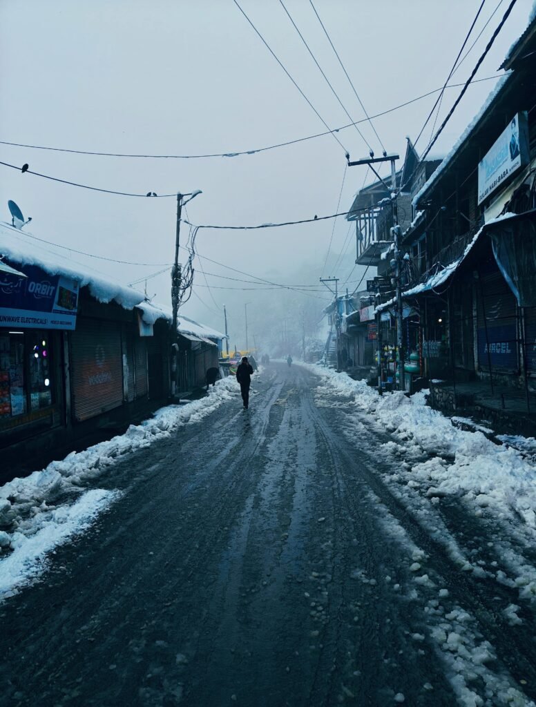 A lone person walks down a foggy, snow-covered street in Charar-i-Sharief during winter.