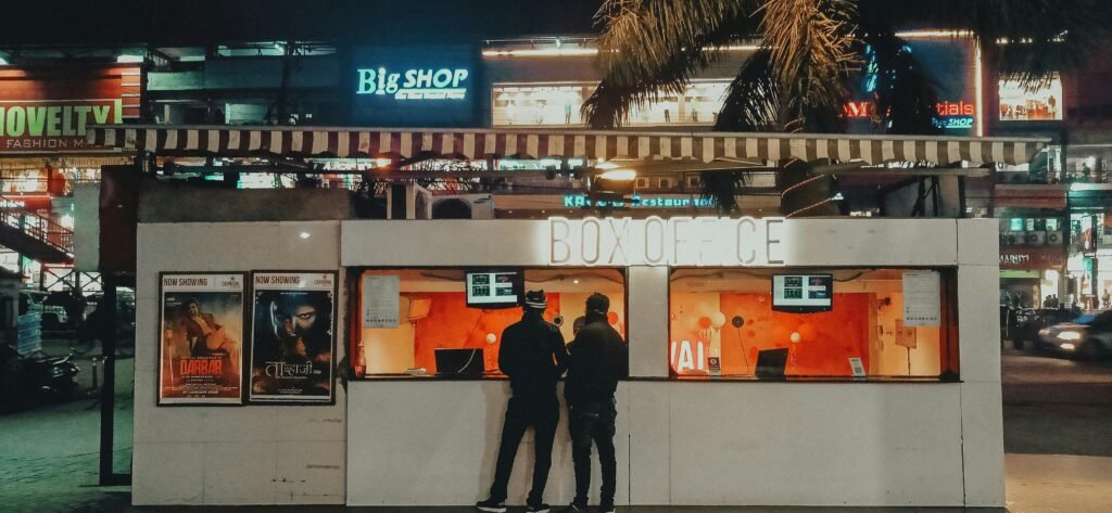Two people standing at a box office under neon lights at night, urban cinema setting.